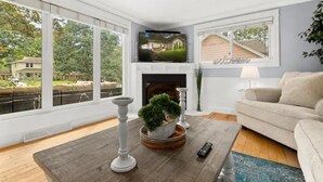 A living room with a fireplace, a television mounted above it, and a large window offering a view of the outdoors.
