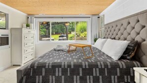 A bedroom with a large bed, a wooden ceiling, a window with a view of greenery, and a white dresser.
