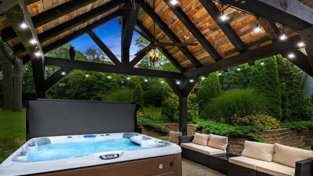 A hot tub under a wooden pergola with string lights, surrounded by a garden and outdoor seating.