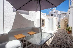 A narrow outdoor corridor with a glass table and two cushions, a patterned rug, and a potted plant.