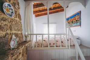 Une chambre avec un mur en pierre, un lit recouvert d’une literie à motifs floraux, un baldaquin blanc et un tableau représentant un paysage.