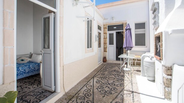 Un couloir étroit avec un sol à motifs, un lit recouvert d’une couverture bleue et blanche, et une petite table sur laquelle est posé un parapluie violet.