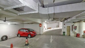 Un estacionamiento con un coche rojo aparcado en un lugar, flechas direccionales en el suelo y un cartel de 'prohibido el paso' en la pared.