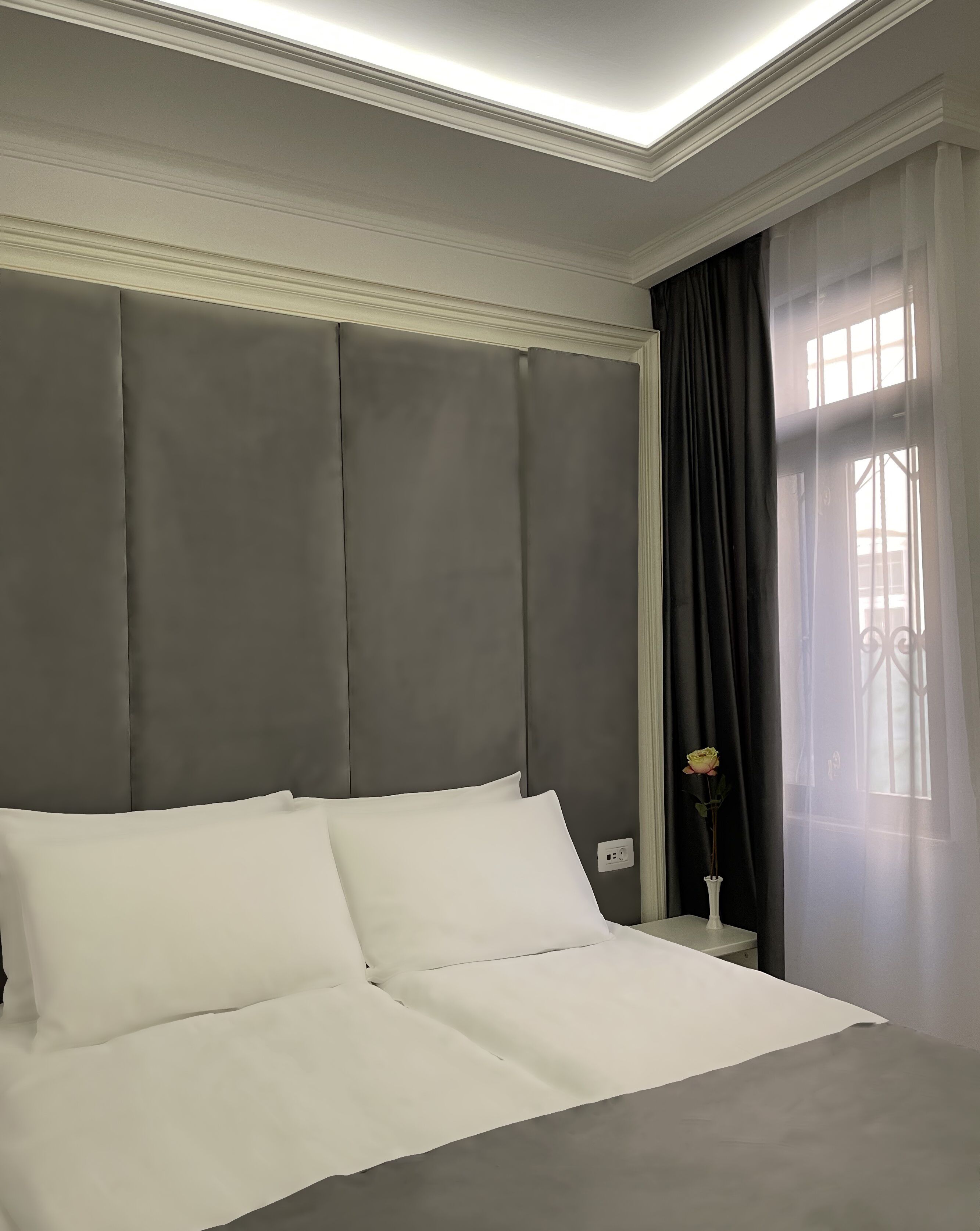 A neatly made bed with white linens in a room with grey padded headboard and a window with sheer curtains.
