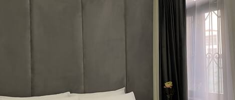 A neatly made bed with white linens in a room with grey padded headboard and a window with sheer curtains.