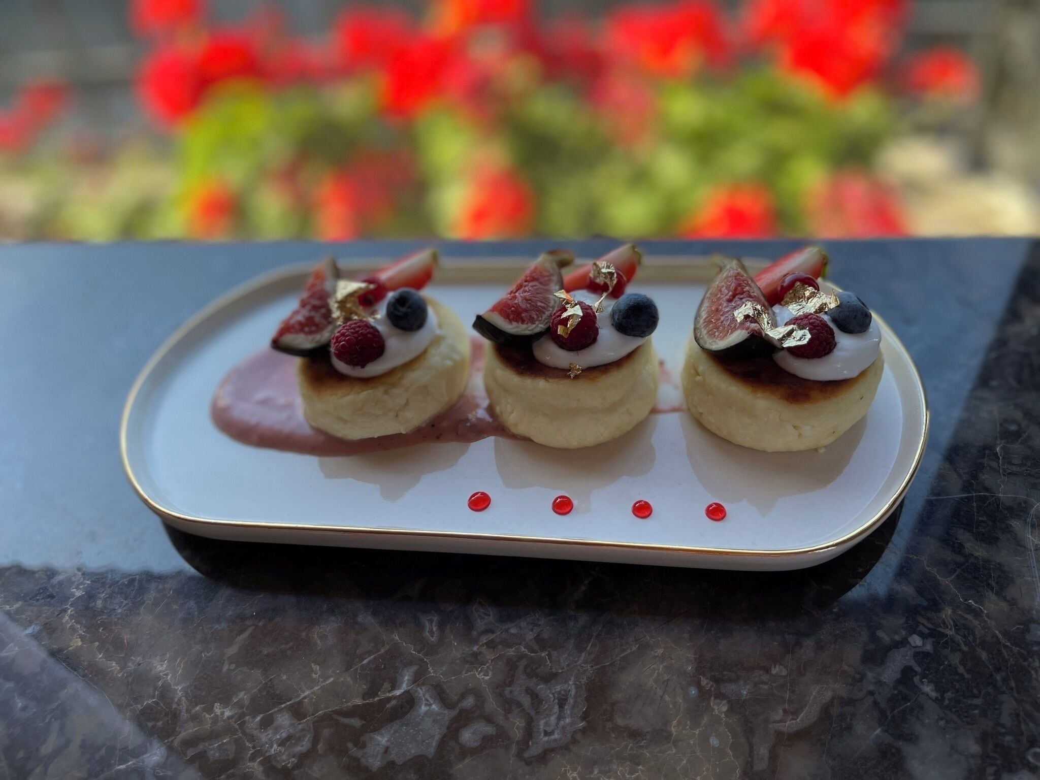 Drei Desserts auf einem weißen Teller mit goldener Kante, auf einer dunklen Marmorplatte platziert.