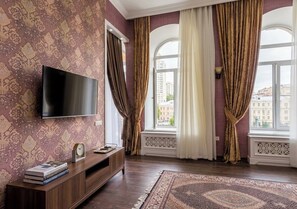A room with a flat-screen TV, a wooden entertainment unit, patterned wallpaper, large windows with curtains, and a patterned rug. - Seven Palace Hotel (Baku)