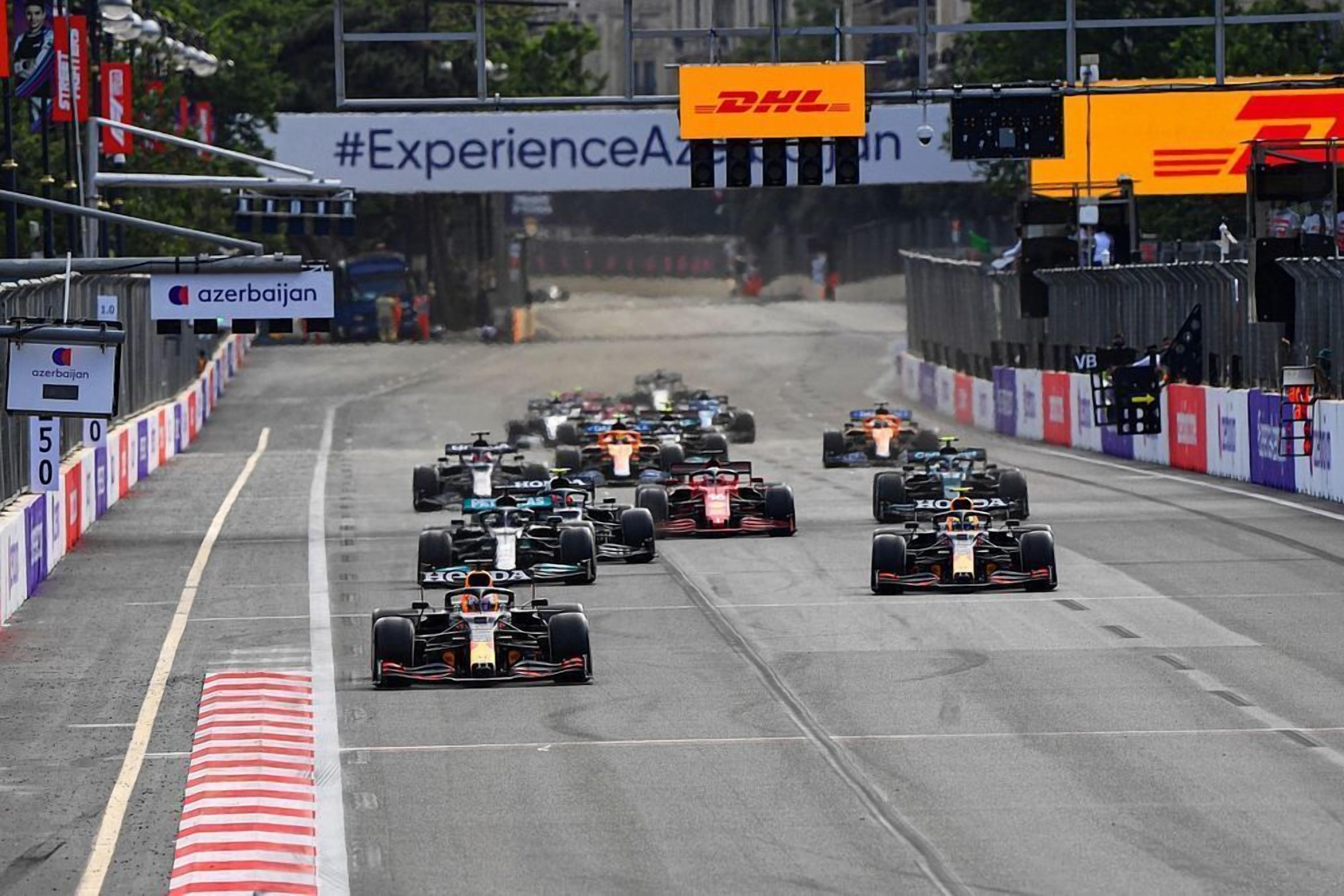 A race track with multiple racing cars lined up at the starting line, surrounded by barriers and sponsor banners.