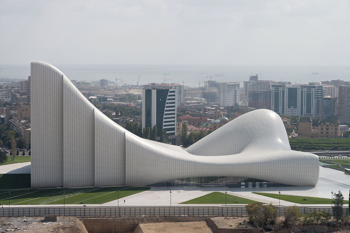Uma estrutura arquitetônica moderna com design ondulado, diante de um horizonte urbano com edifícios altos e uma vista distante do mar.