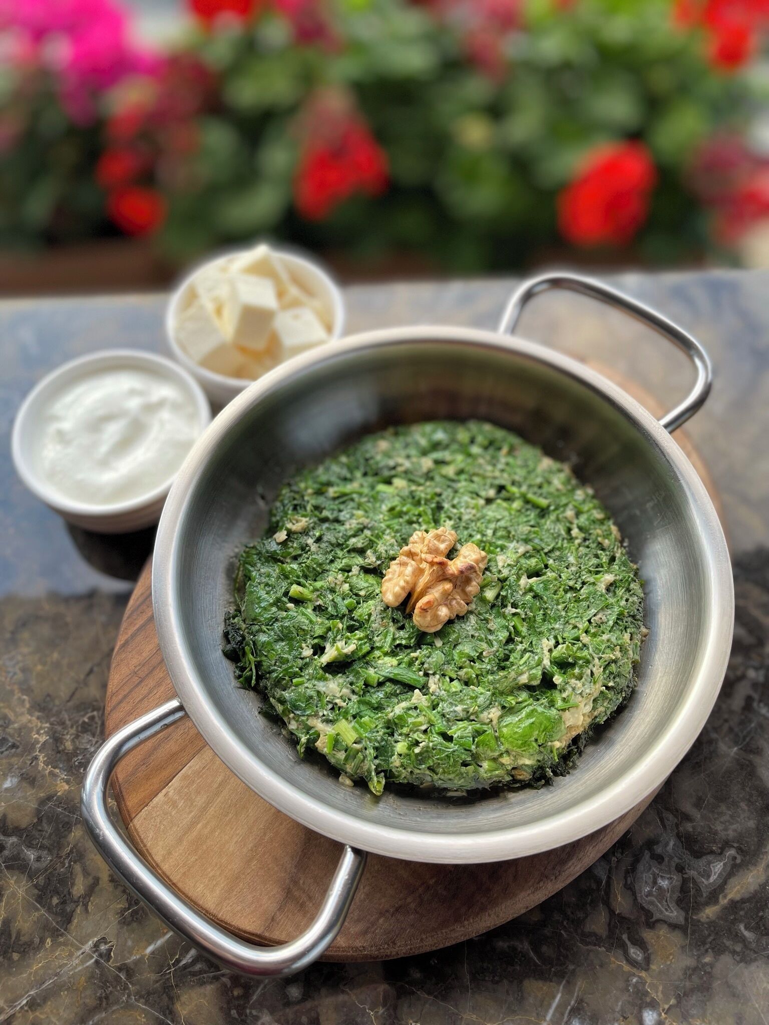 A metal bowl with a wooden base containing a green dish, accompanied by small bowls of white and yellow sauces.