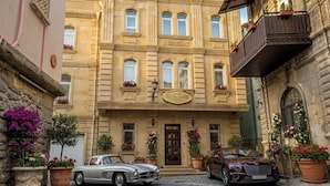 Ein historisches Gebäude mit einem zentralen Eingang, einem Balkon auf der rechten Seite und zwei geparkten Autos davor.