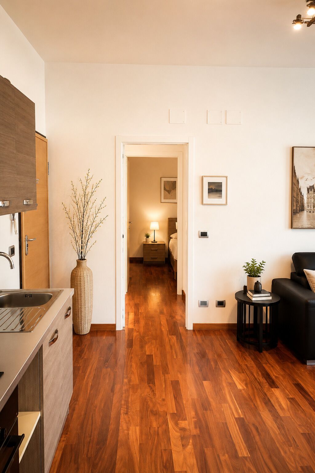 Ein modernes Interieur mit Holzboden, einer Küchenzeile, einem Wohnzimmer mit schwarzem Sofa und einem Flur, der zum Schlafzimmer mit Nachttischlampe führt.
