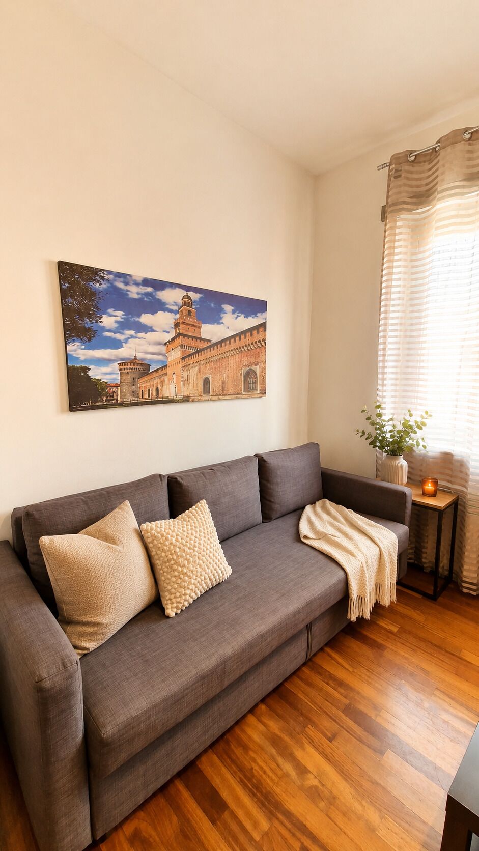 A living room with a grey sofa, a painting of a building, a small table with a plant, and a window with curtains.