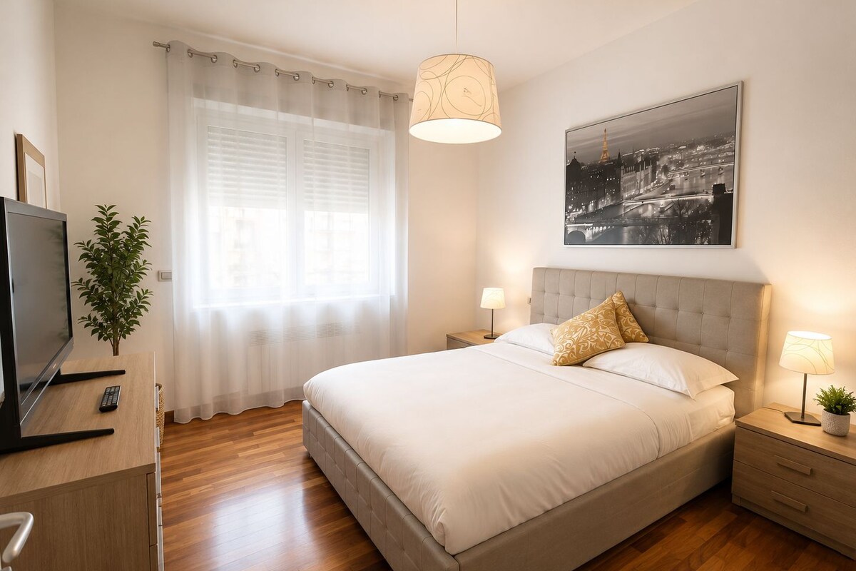 Une chambre à coucher avec un lit, des tables de chevet, une télévision, une fenêtre avec des rideaux, une plante et un tableau représentant un paysage urbain accroché au mur.