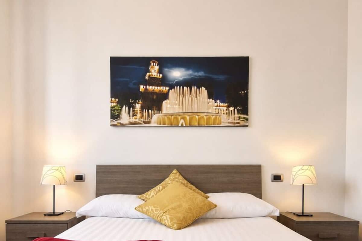 A hotel room with a large bed, two bedside tables with lamps, a nightstand, a red throw pillow, and a framed picture of a cityscape.