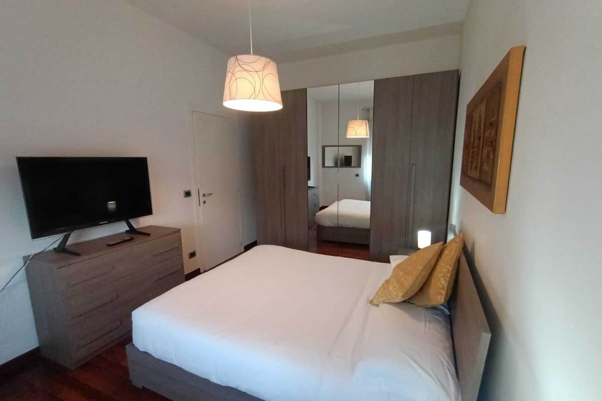 A bedroom with a bed, a TV on a wooden dresser, a mirror, and a hanging light fixture.