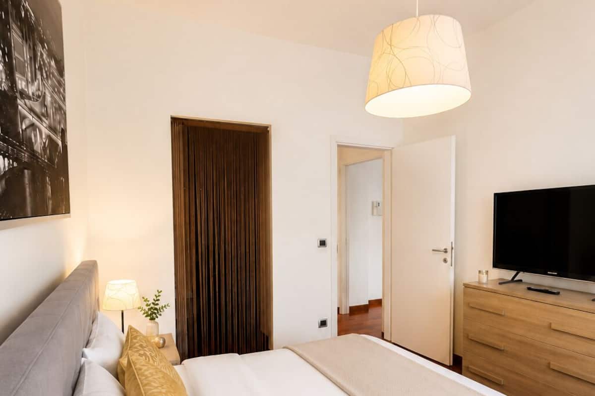 A neatly made bed with white linens, a grey headboard, and a beige throw blanket. There is a wooden dresser with a flat-screen TV on top. A large window with a view of a building is visible in the background.