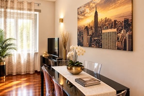 A modern living room with a cityscape painting, a flat-screen TV, a dining table with chairs, and a potted plant. - Style Homes Brera (Milan)