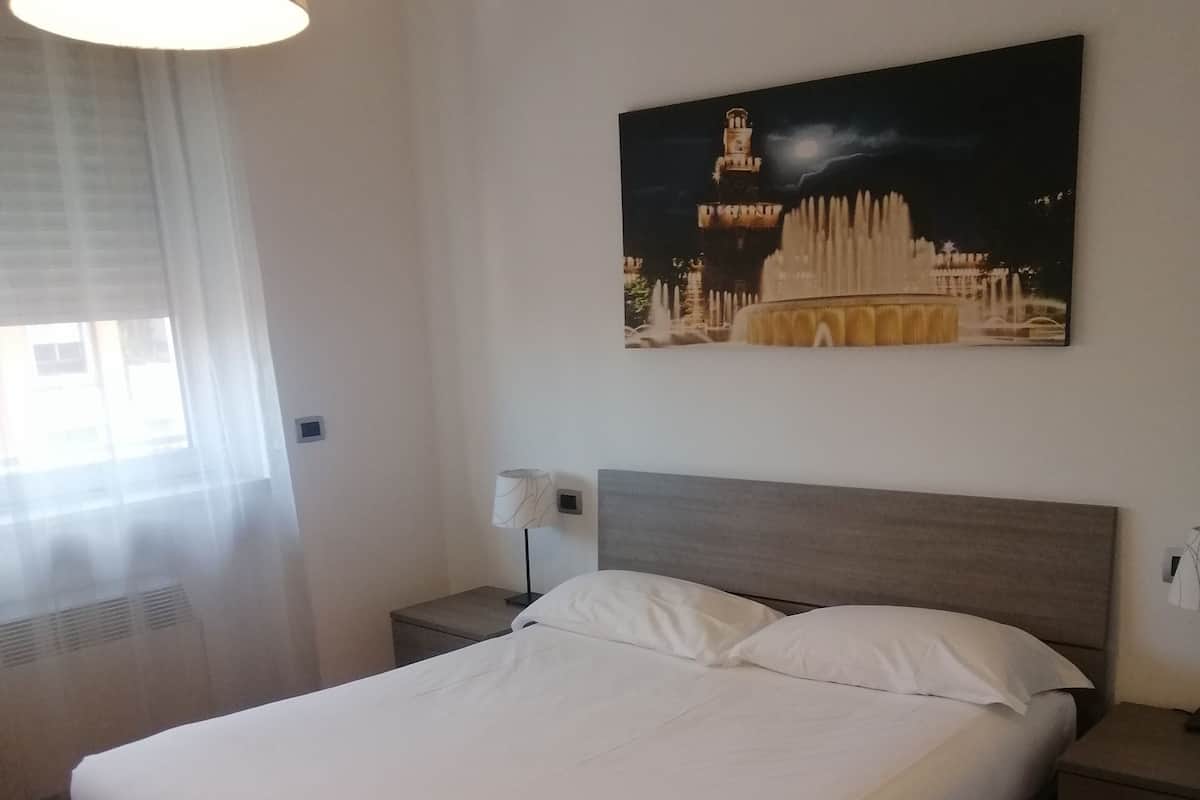 A neatly made bed in a room with a large window, a wall-mounted picture, and a bedside lamp.