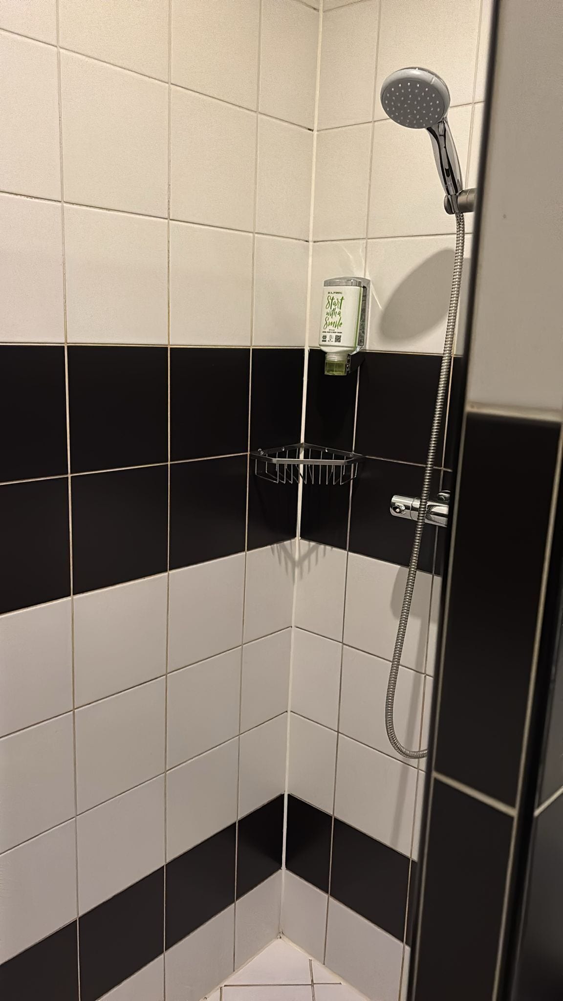 A corner of a bathroom with black and white tiles, a shower head, and a bottle of mouthwash on a shelf.