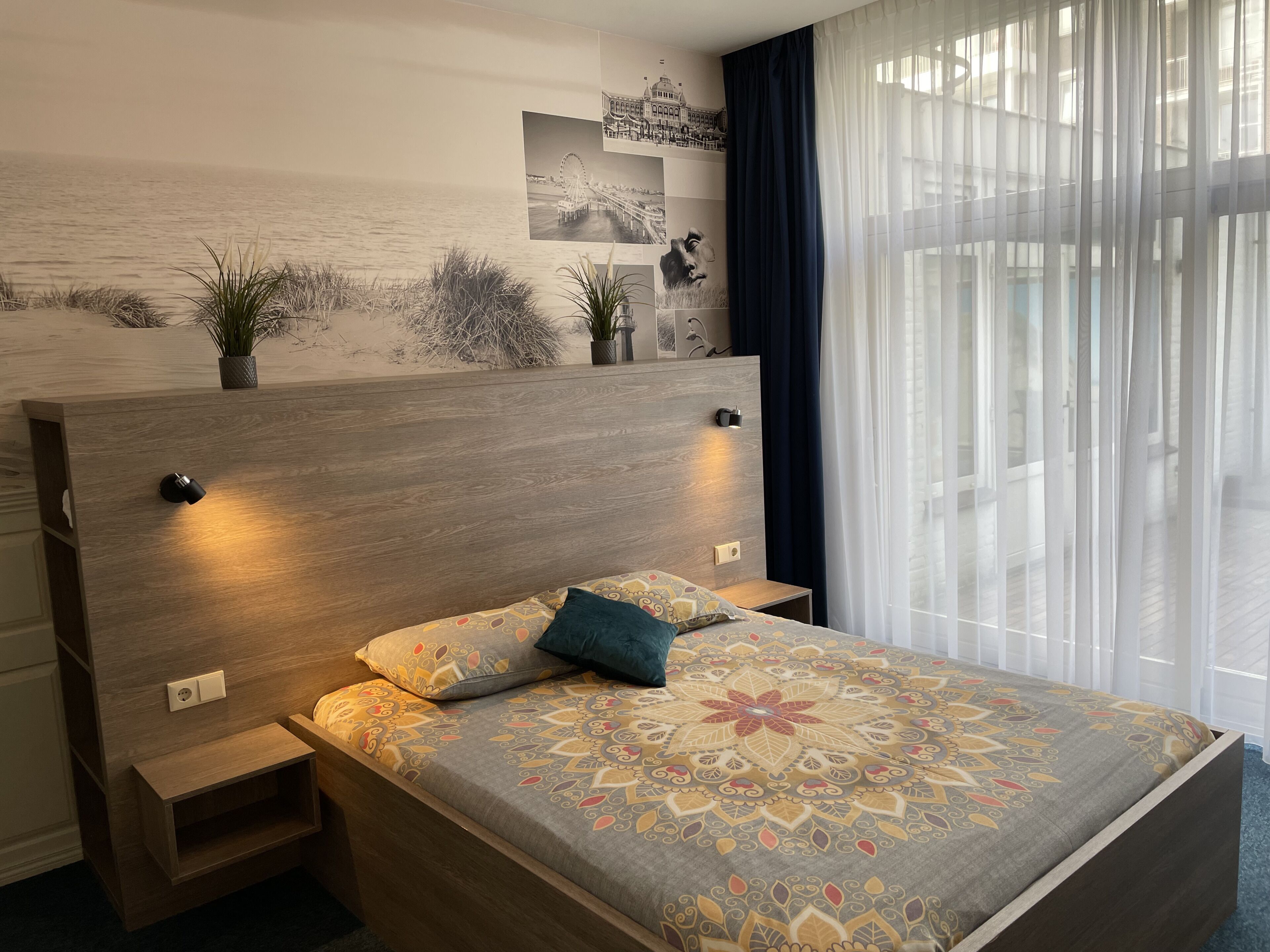 A bedroom with a bed, bedside tables, a headboard with built-in lighting, a wall-mounted artwork, and a view through sheer curtains.