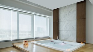A modern bathroom with a large bathtub, wooden flooring, a marble wall, and a city view through large windows. - Riviera Rayhaan by Rotana Doha (Doha)