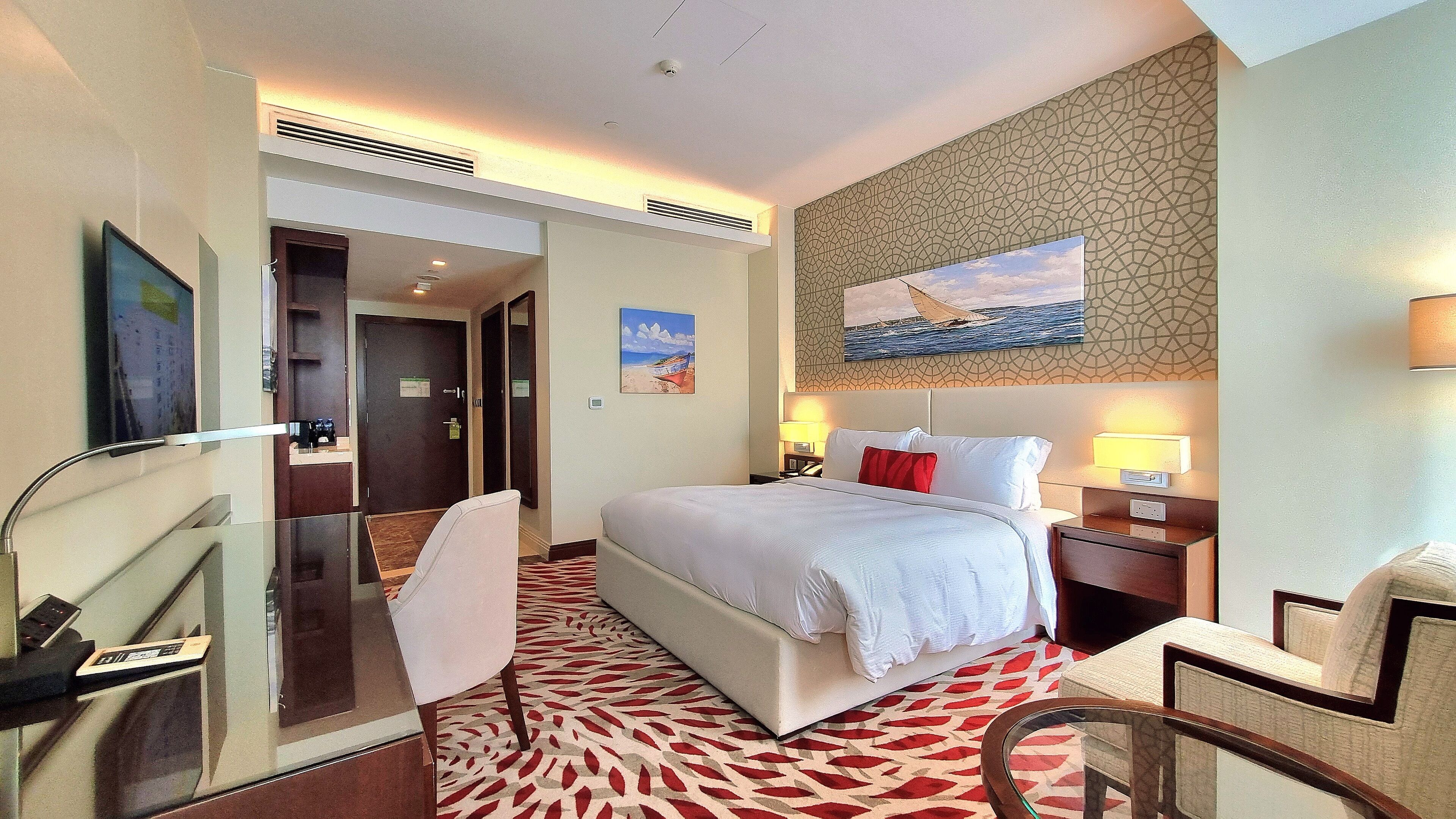 A hotel room with a bed, a desk, a chair, a painting on the wall, and a patterned carpet.