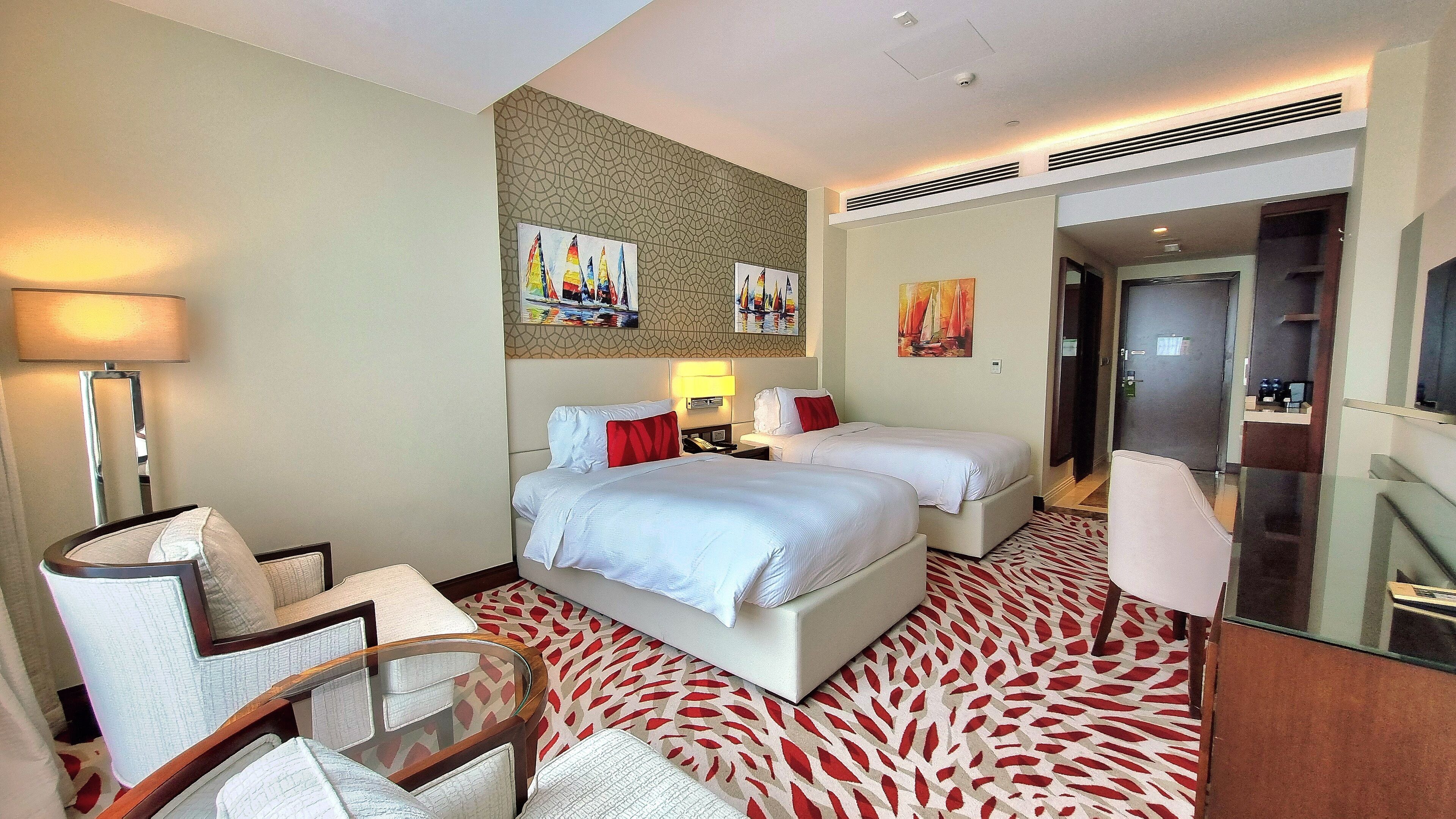 A hotel room with two beds, a sitting area with a chair, a desk, and a lamp. There are paintings on the wall and a door leading to a bathroom.