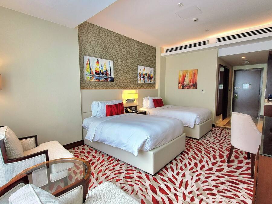 Ein Hotelzimmer mit zwei Betten, einer Sitzecke mit einem Sessel, einem Schreibtisch und einer Lampe. An den Wänden befinden sich Gemälde und eine Tür führt ins Badezimmer.