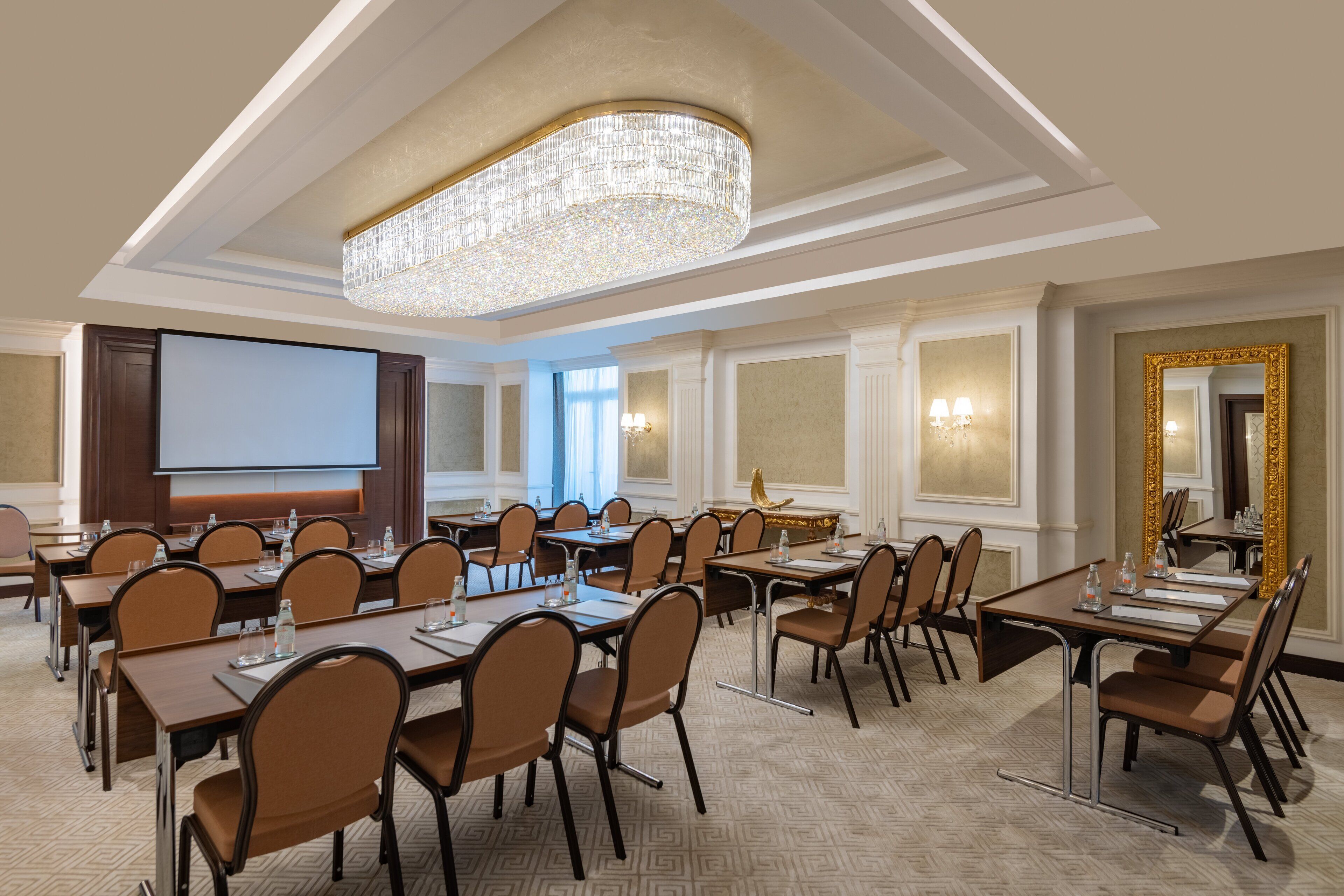 A conference room with a large screen, multiple tables, and chairs, featuring a chandelier and elegant wall decor.