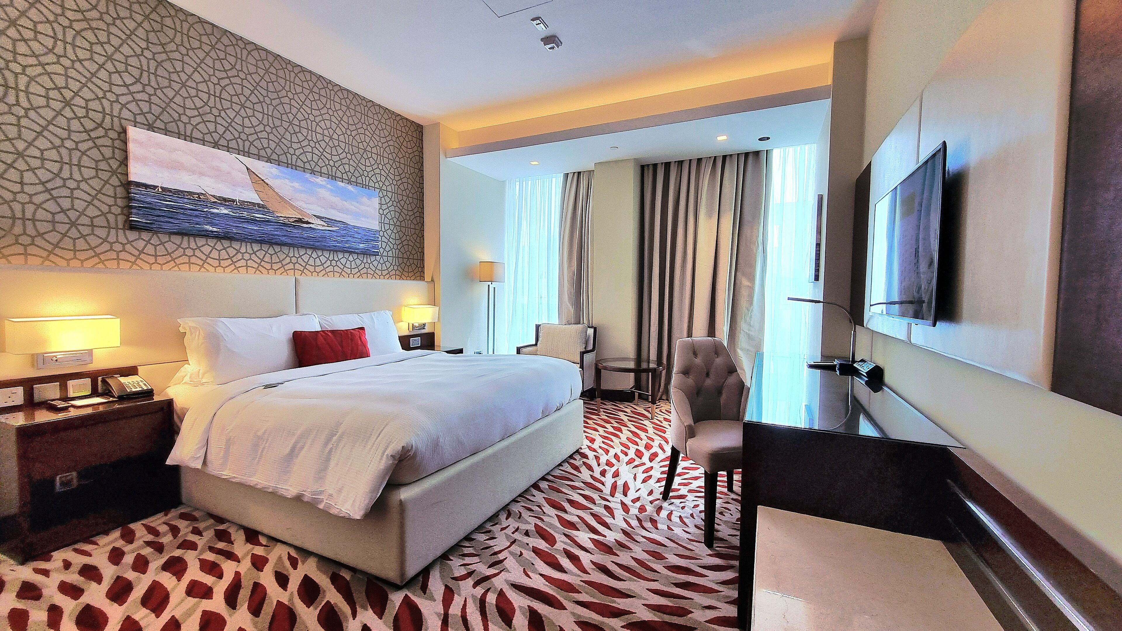 A hotel room with a large bed, a desk with a flat-screen TV, a chair, a desk lamp, and a painting on the wall.