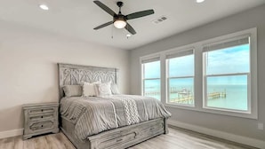 A bedroom with a bed, nightstands, a ceiling fan, large windows, and a view of the ocean. - Soggy Pollards (Rockport)