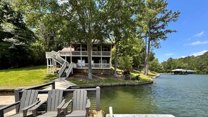 A lakeside dock with Adirondack chairs, a wooden deck, and a house with a balcony and stairs leading to a deck.