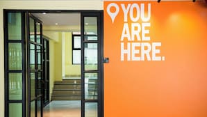 Een binnengang met glazen deuren die leiden naar een trap, en een oranje muur met een markering voor een locatie.