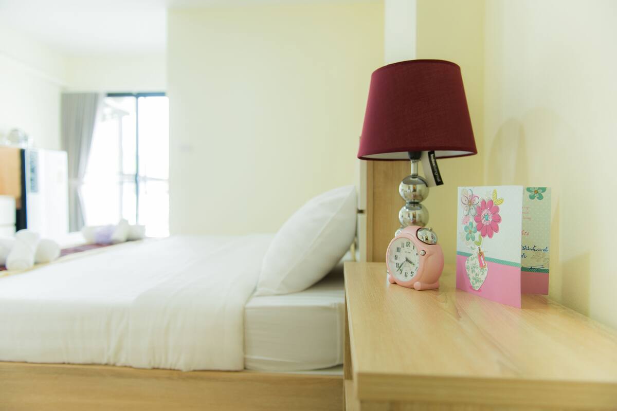 A neatly made bed with white bedding, a wooden nightstand with a red lampshade, a pink alarm clock, and a card with floral design.