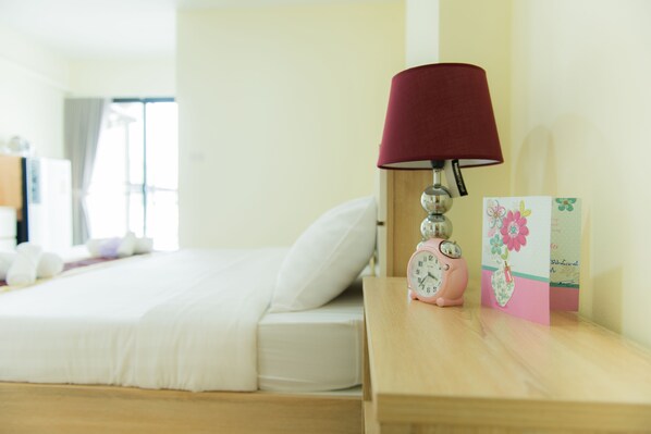A neatly made bed with white bedding, a wooden nightstand with a red lampshade, a pink alarm clock, and a card with floral design. - Be my guest36 (Bangkok)