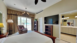 A spacious bedroom with a large bed, a sitting area with a chair, a television, and a bathroom with a vanity. - En Soleil #121 West | Gulf Front, Large Patio + Amenities (Panama City Beach)