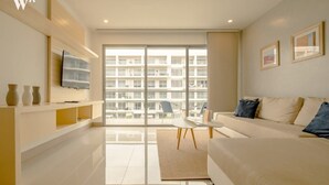 A modern living room with a sofa, a TV mounted on the wall, and a large window offering a view of a building. - w* | Stunning 1BR with Pool in Cartagena (Cartagena de Indias)