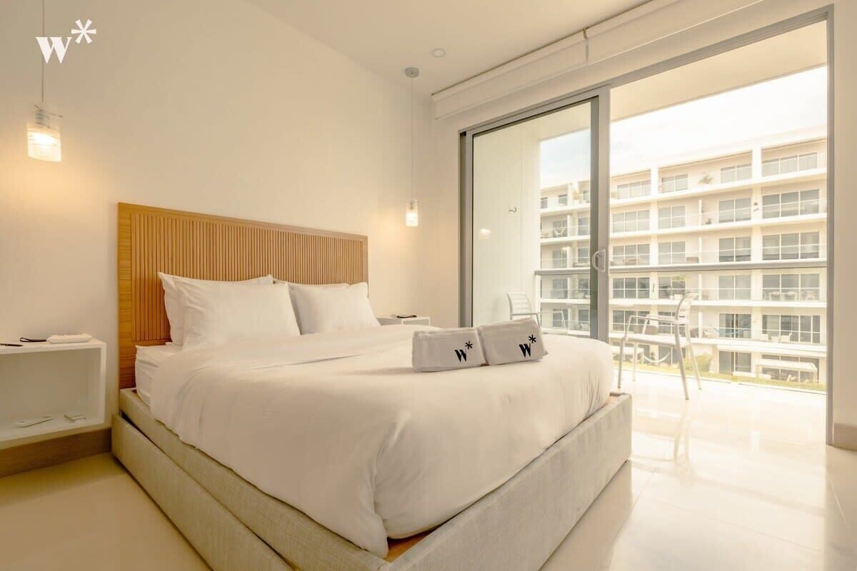 Una habitación de hotel moderna con una cama grande, cabecera de madera, una mesita de noche y un balcón con vistas a otros edificios.