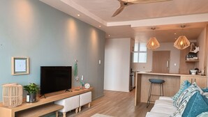 Una sala de estar moderna con televisor de pantalla plana, muebles de madera, ventilador de techo y una zona de cocina con taburetes de bar.