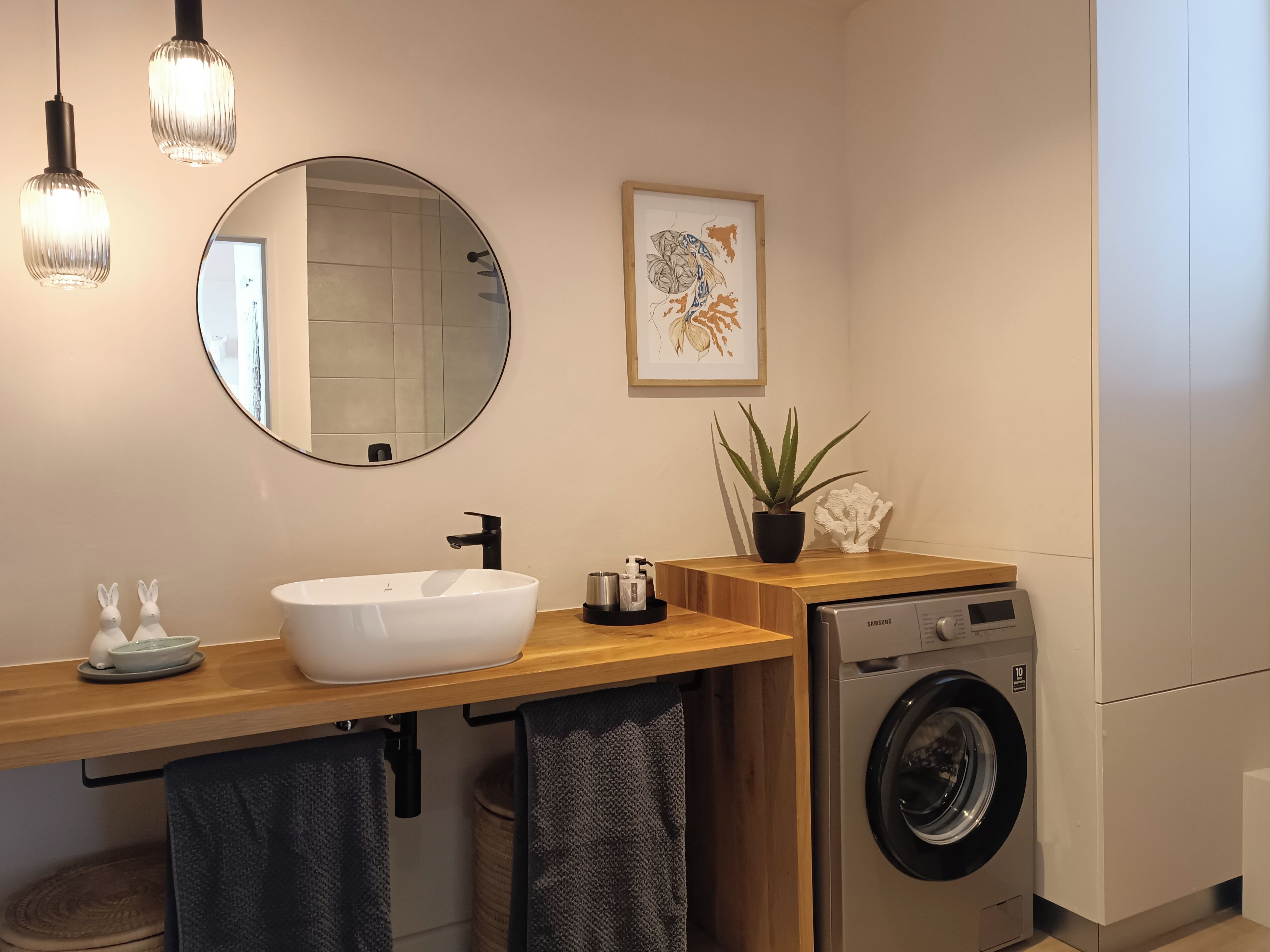 A modern bathroom with a white sink, black faucet, round mirror, and a Samsung washing machine.