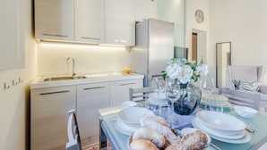 A modern kitchen with white cabinets, a stainless steel refrigerator, and a dining area set with white plates, glasses, and bread.