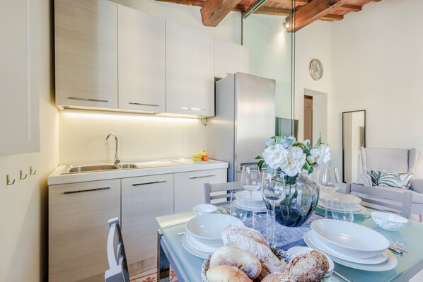 A modern kitchen with white cabinets, a stainless steel refrigerator, and a dining area set with white plates, glasses, and bread.