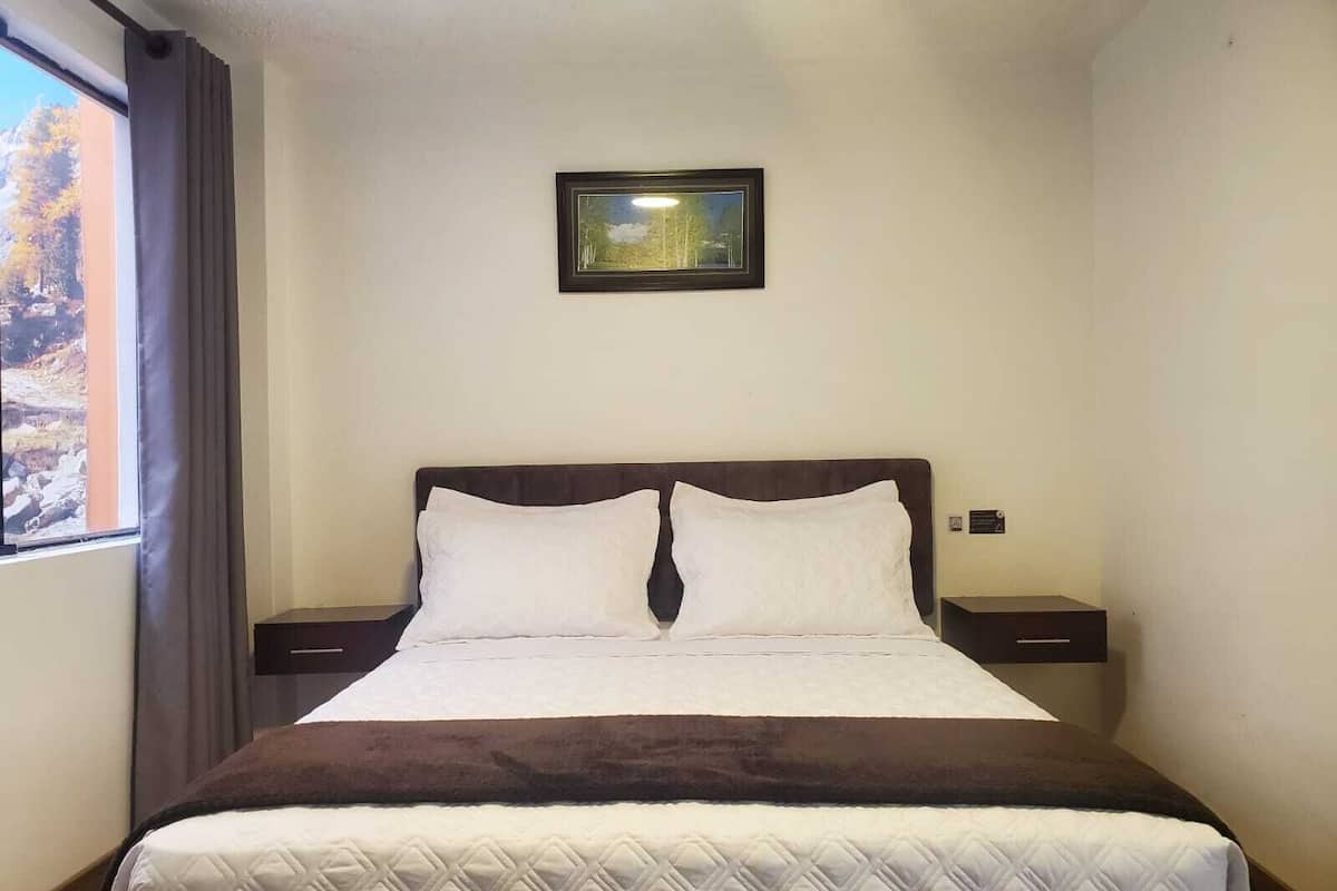 A neatly made bed with a quilted cover, two bedside tables with lamps, a framed picture on the wall, and a window with curtains.