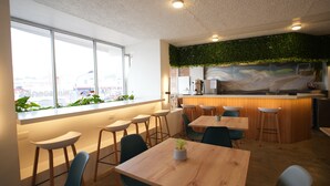 Ein modernes Café mit Barbereich, Holztischen und -stühlen, großen Fenstern mit Stadtblick und einer begrünten Wand mit Pflanzen.