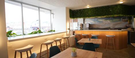 A modern cafe with a bar area, wooden tables, and chairs, large windows with a view of the city, and a green wall with plants.