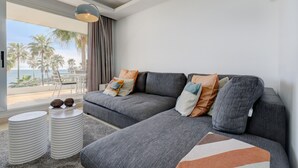 Un salon moderne avec un canapé d’angle gris, des petites tables d’appoint rondes blanches et une vue sur la plage.