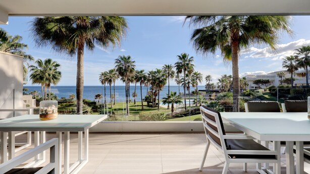 Un balcon avec vue sur la mer, des palmiers, et une table blanche avec une chaise.