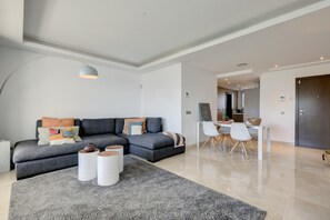 A modern living room with a sectional sofa, a dining area with a white table and chairs, and a kitchen in the background.