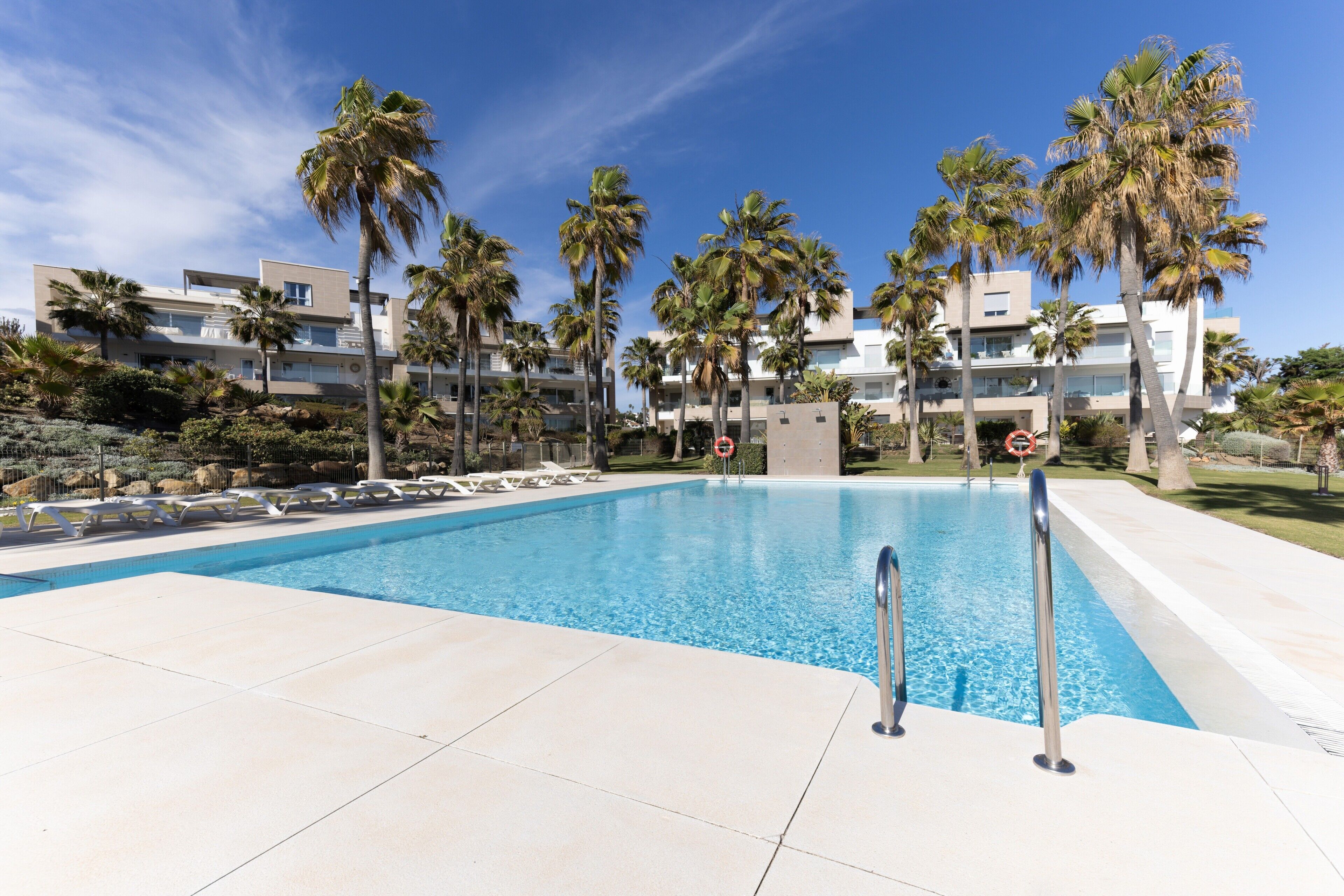 Una piscina con sillones reclinables, palmeras y edificios modernos al fondo.